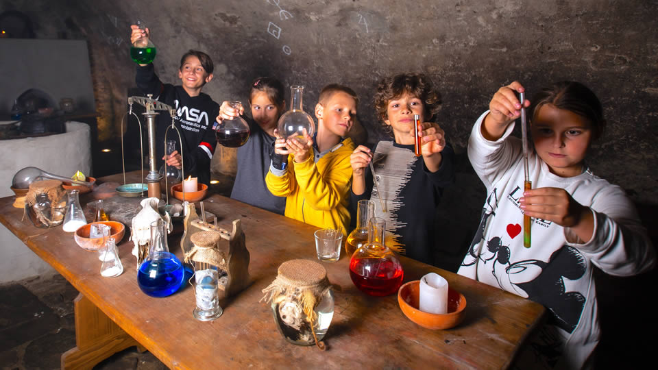 Dětská interaktivní dílna v Klášterech Český Krumlov. Děti stojí okolo stolu při dělání různých chemických pokusů.